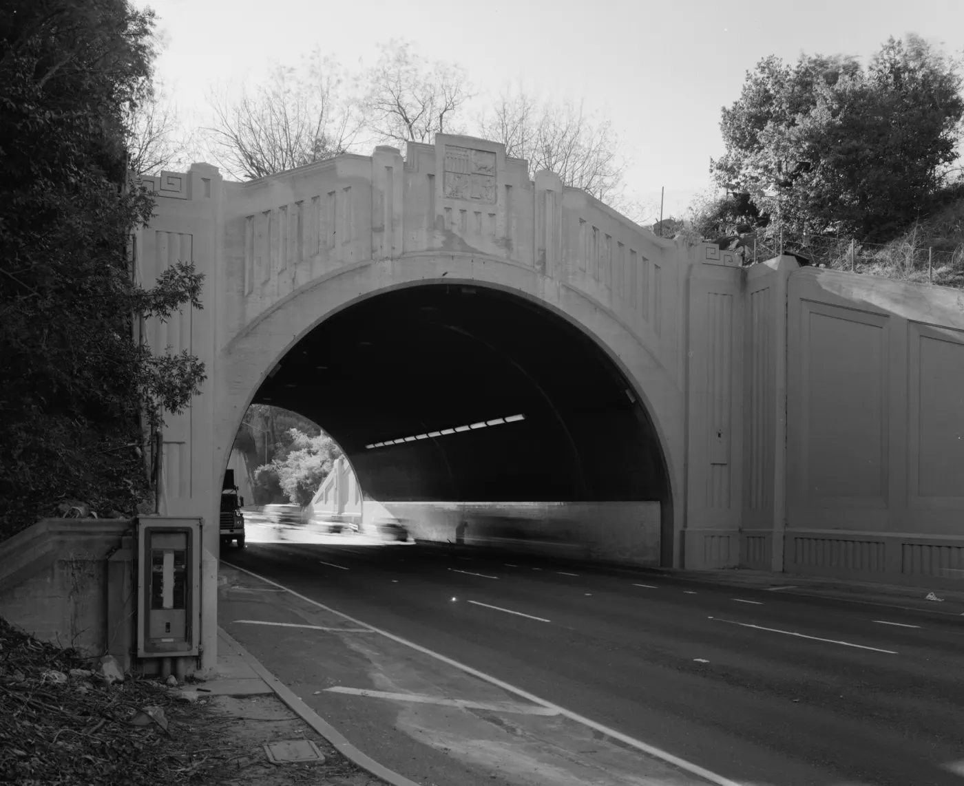 A black and white photograph of a cast concrete Art Deco tunnel portal, with a carved panel at the crown of the arch, a strip of lights running away inside the bore, and a vehicle blurred by a long exposure at the far end.
