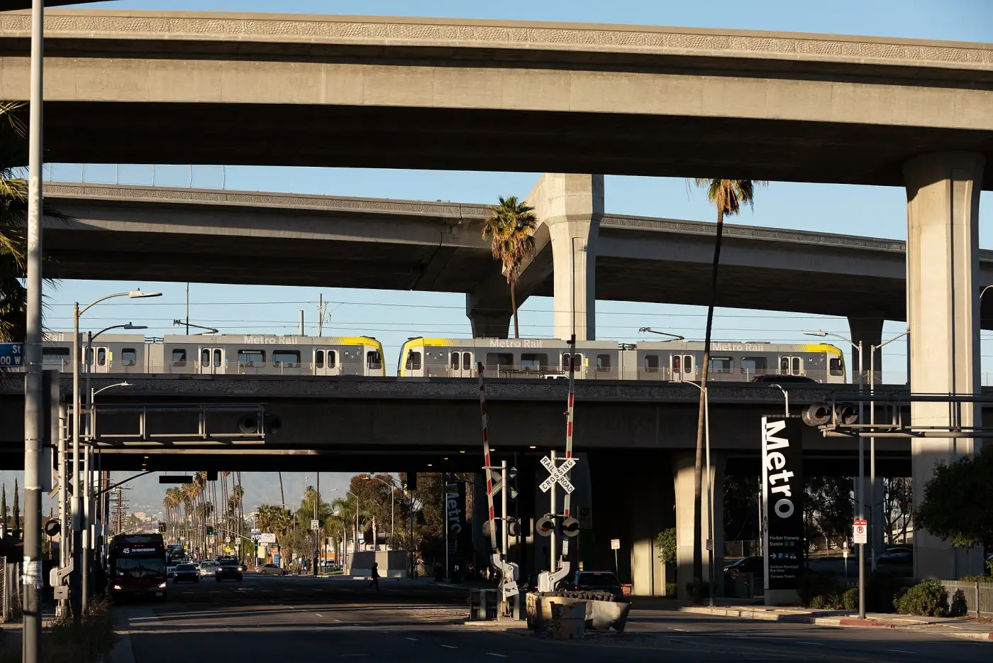 Stacked concrete freeway decks carried on single columns, with a Metro Rail train on its own elevated deck below them and a Harbor Freeway Station sign at street level.