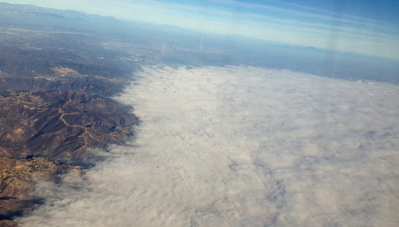 An aerial view of a flat white cloud deck filling the lower half of the frame and ending in a ragged edge against dark mountain ridges, with dry country and a distant range beyond it.