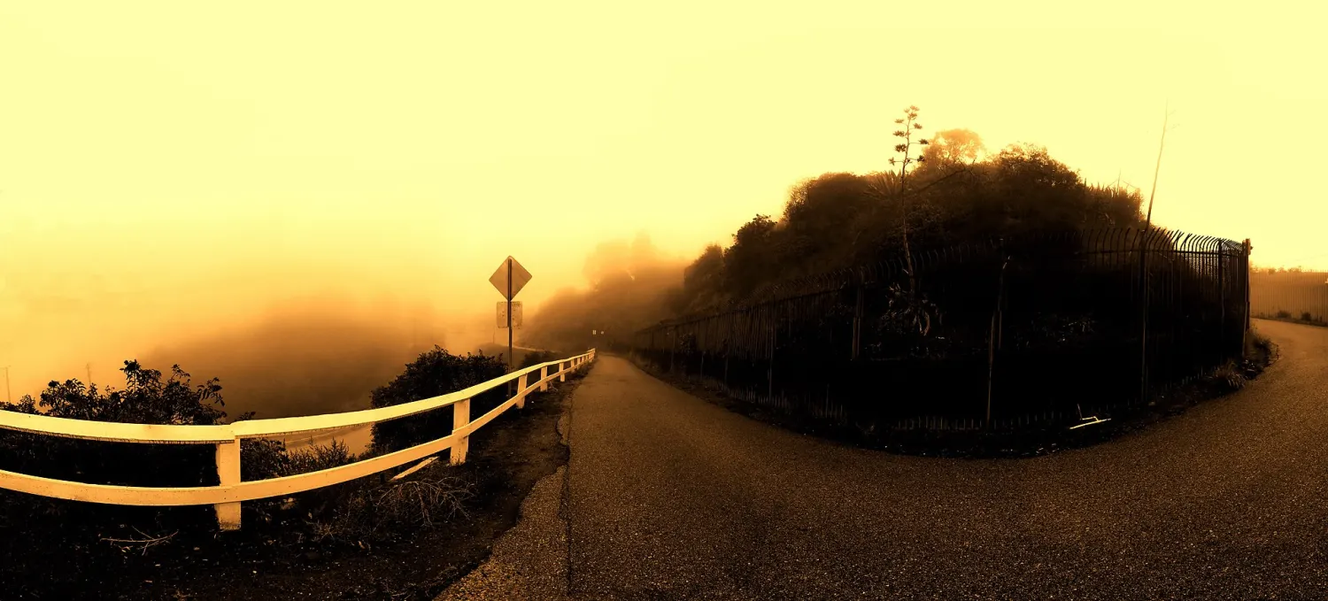 A ridge road running into fog, with a white guardrail on the left above a bank of cloud and a fenced hillside on the right, the whole scene toned gold.