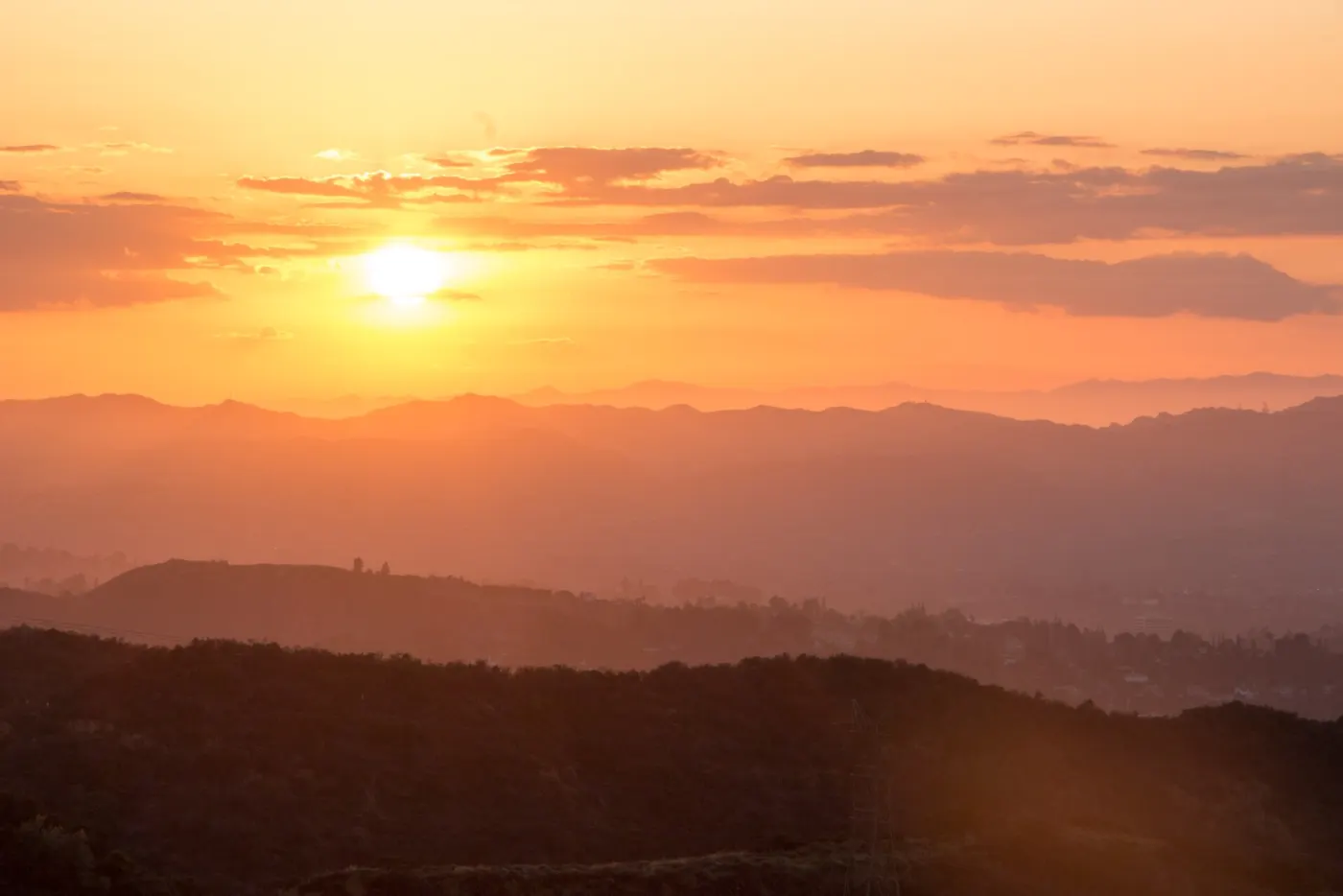 Sunset over layered ridges, with the San Fernando Valley hazy below and the first city lights showing on the right under an orange sky.